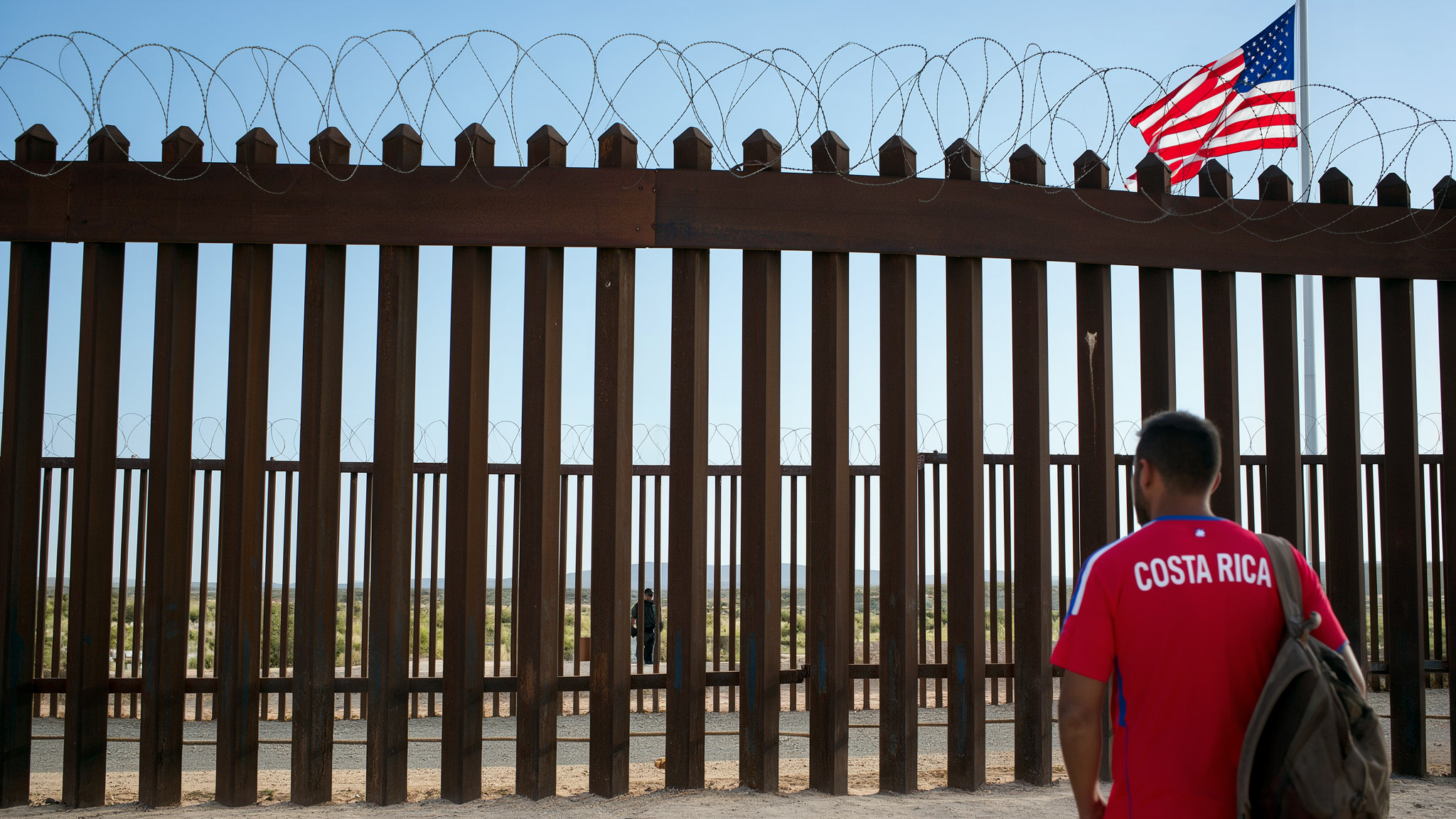 Hombre con camiseta roja de Costa Rica, de espaldas, frente al muro fronterizo de Estados Unidos; detrás del muro, la bandera estadounidense y un agente a lo lejos
